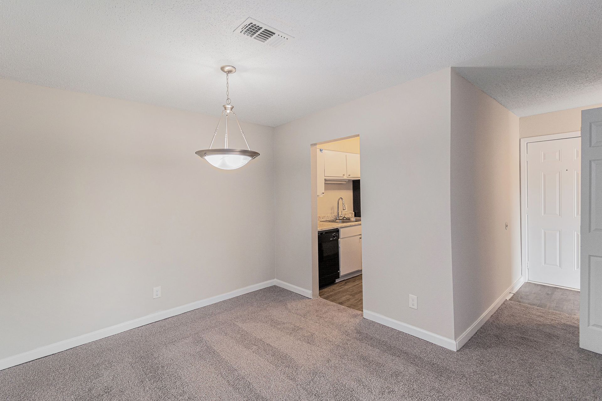 A neutral-colored dining room with grey carpet, a pendant light fixture, and an open doorway leading into a kitchen.