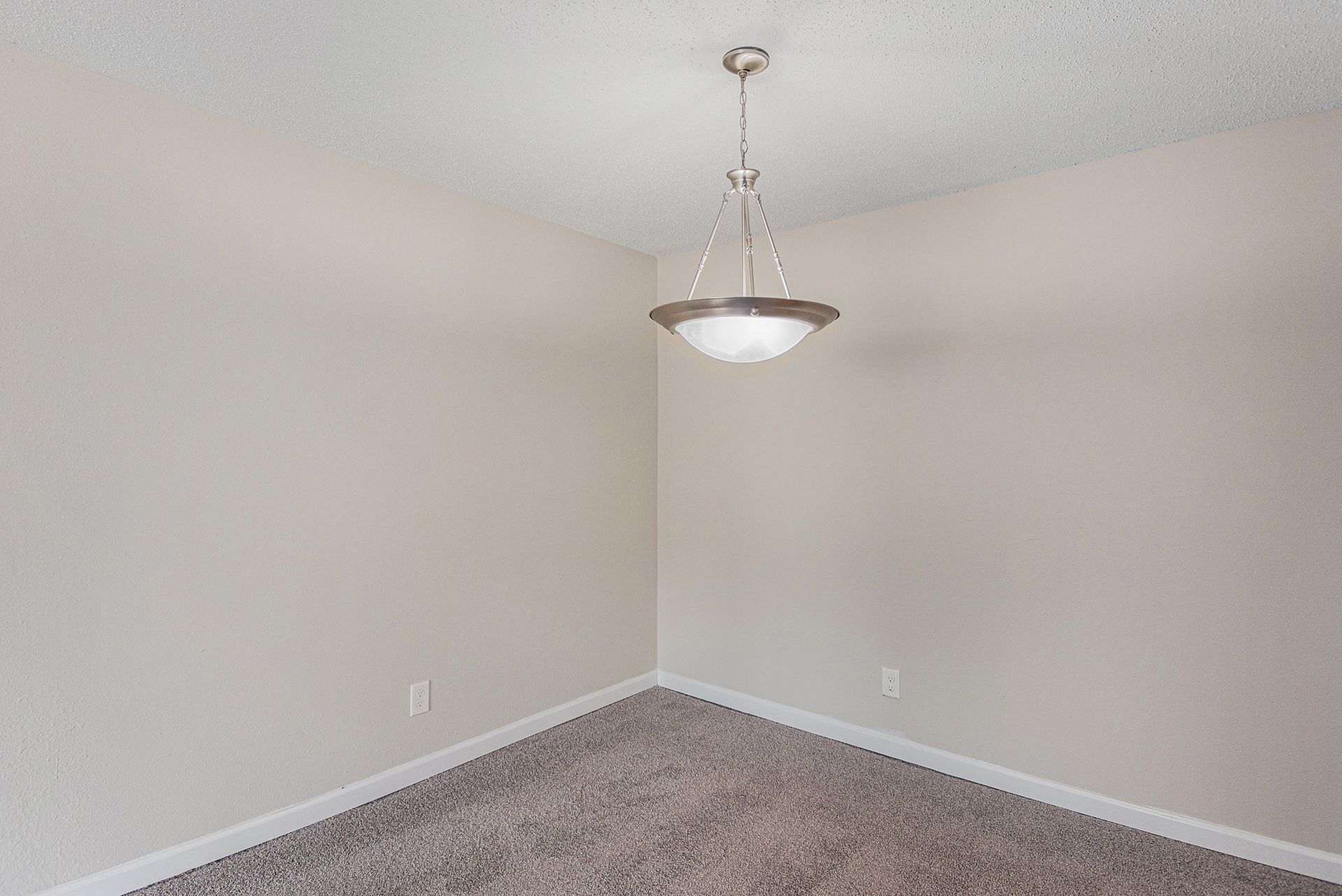 An empty room corner with beige walls, gray textured carpeting, and a modern, tiered pendant light fixture.