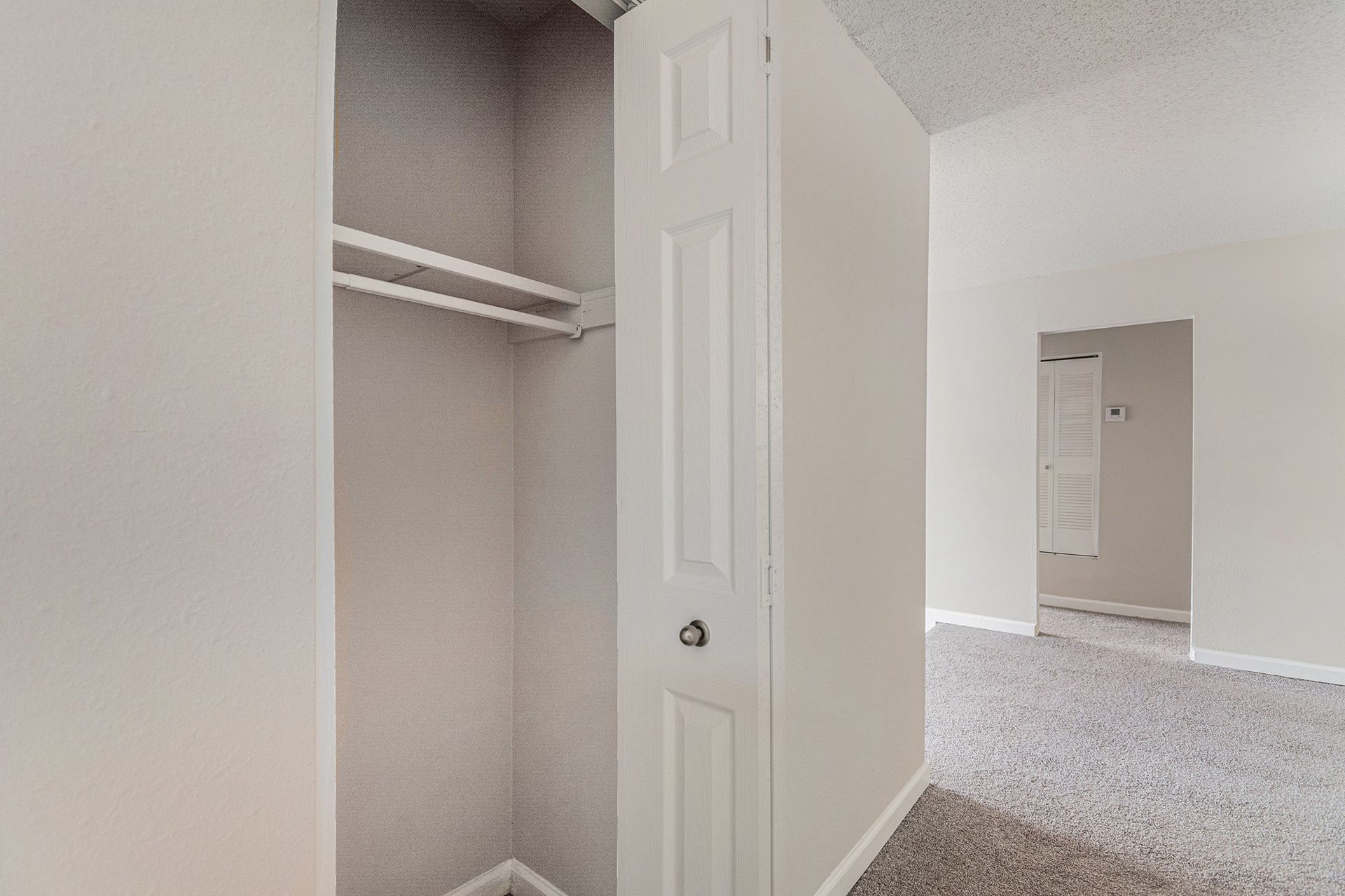 An open white closet with a shelf, set in a room with light grey walls and grey carpet, leading to another doorway.