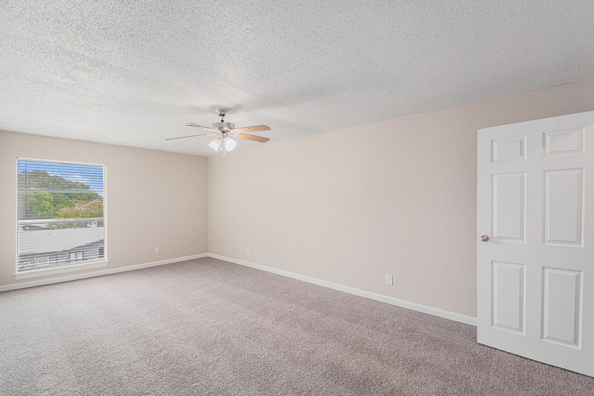 Empty bedroom with beige walls, gray patterned carpet, a ceiling fan, and a white six-panel door.