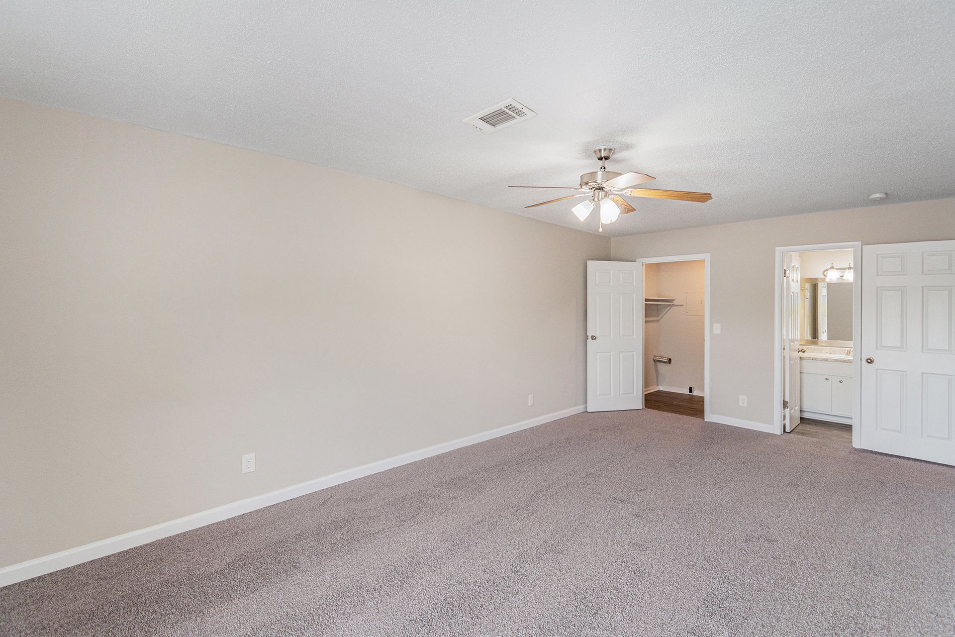 A neutral, carpeted bedroom with white walls, a ceiling fan, a closet door, and a partially open bathroom doorway.