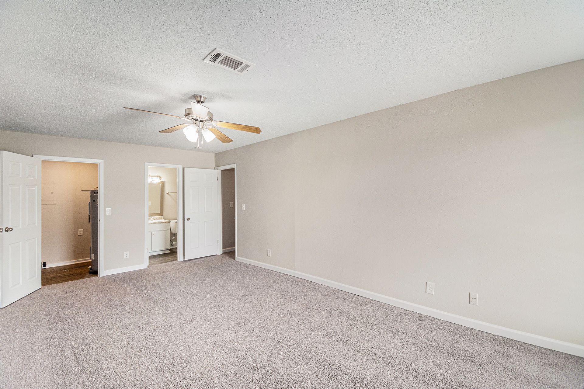 A neutral-colored bedroom featuring gray carpet, a ceiling fan, and open doorways leading to a bathroom and closet.