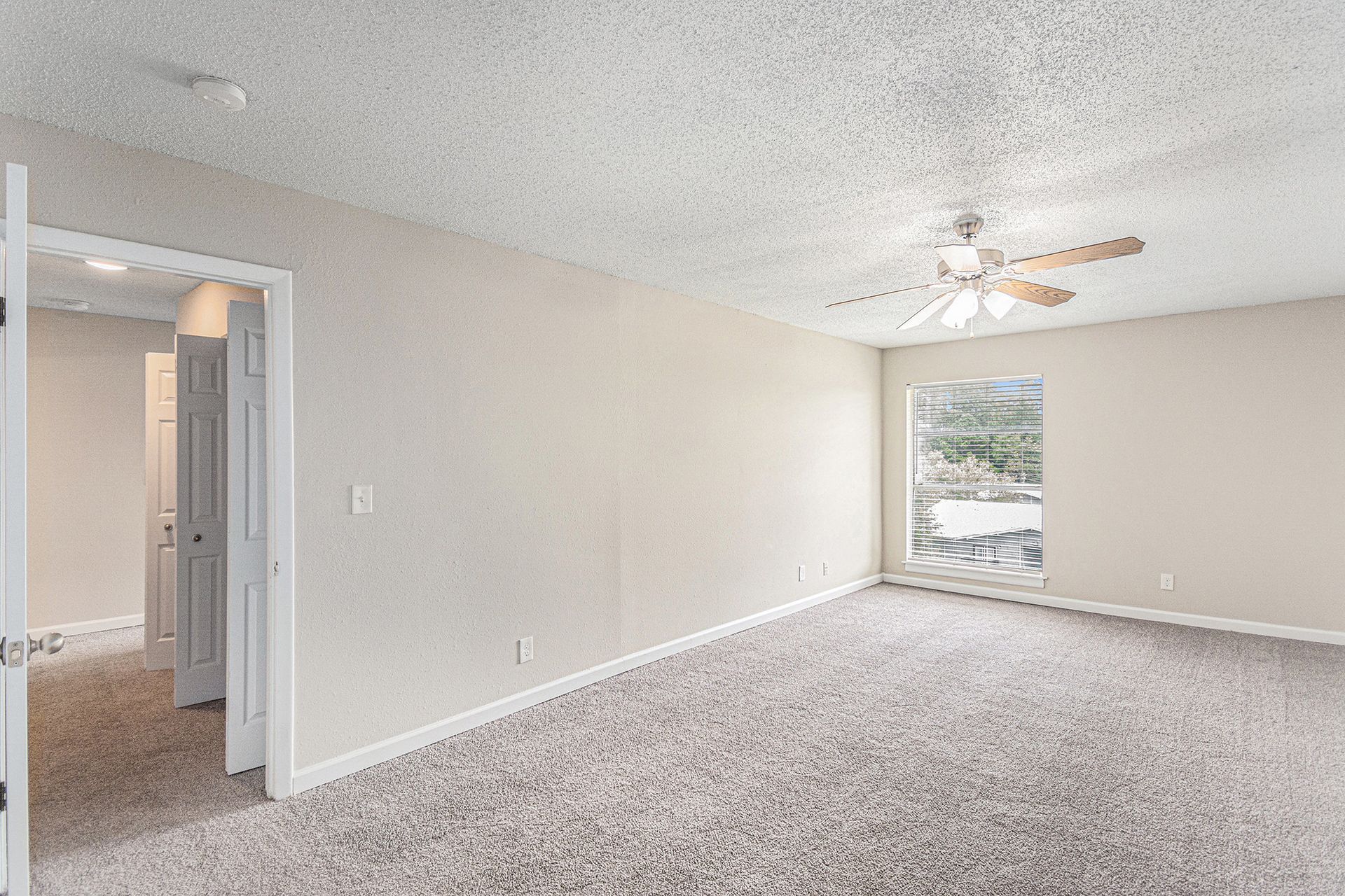 A spacious bedroom with beige walls, patterned carpet, a ceiling fan, and an open doorway leading to a hallway.