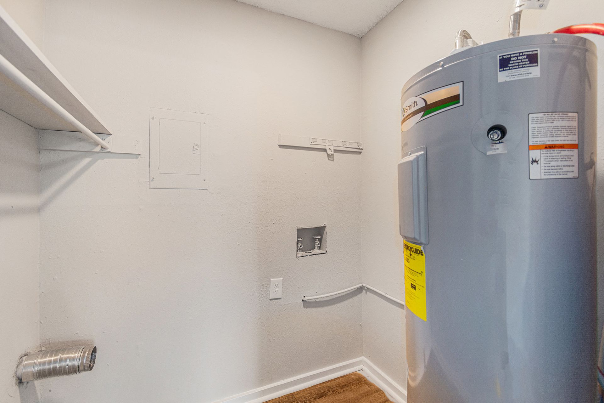 A utility closet featuring a water heater, a washer hookup, and a wall-mounted shelf over a wooden floor.