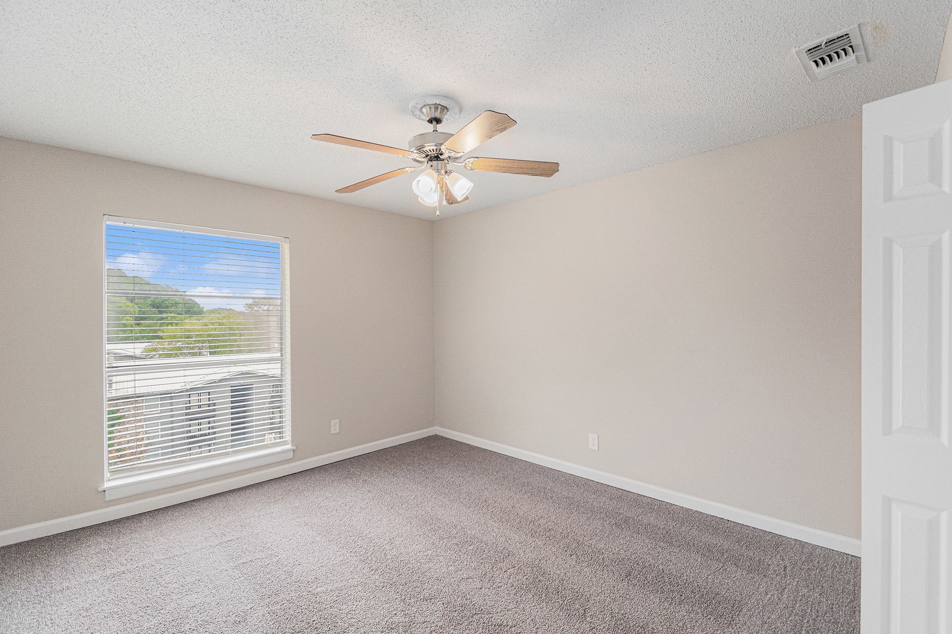 An empty bedroom with beige walls, grey carpet, a ceiling fan, and a window with a view of a neighborhood.