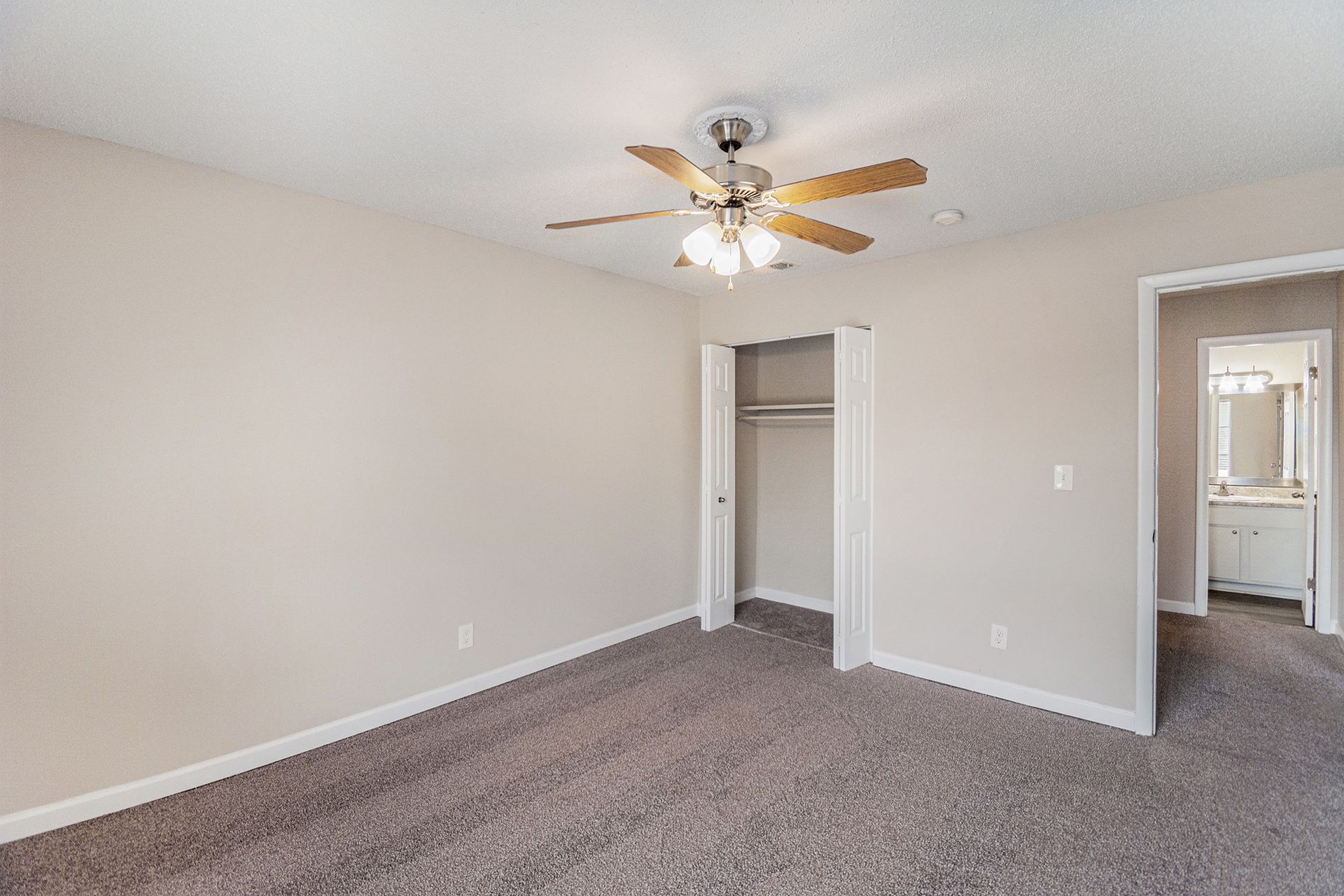 A carpeted bedroom with light beige walls, a ceiling fan, a walk-in closet, and an open doorway leading to a bathroom.