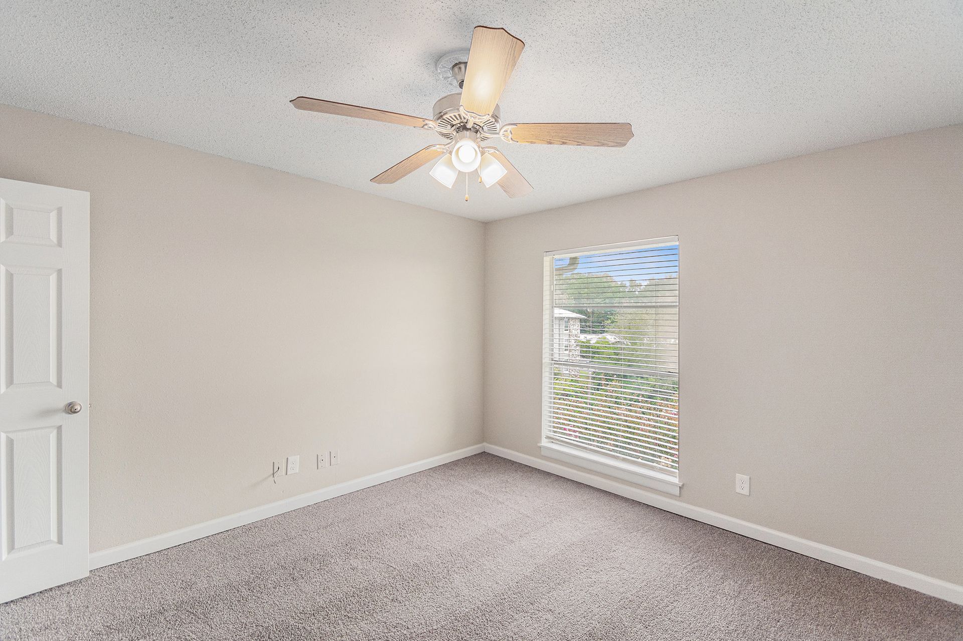 An empty bedroom with light beige walls, gray carpet, a ceiling fan, and a single window with blinds.