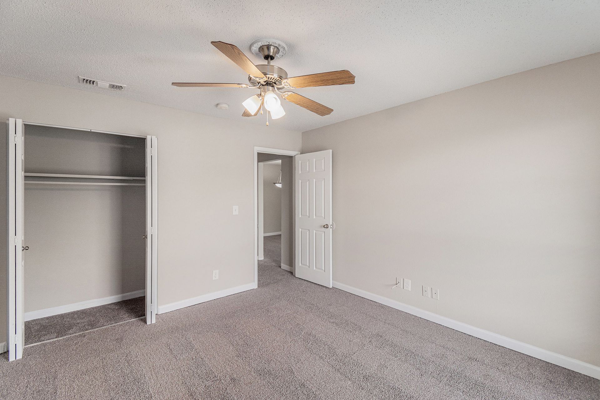 An empty bedroom with gray carpet, beige walls, a ceiling fan, an open closet, and a white interior door.