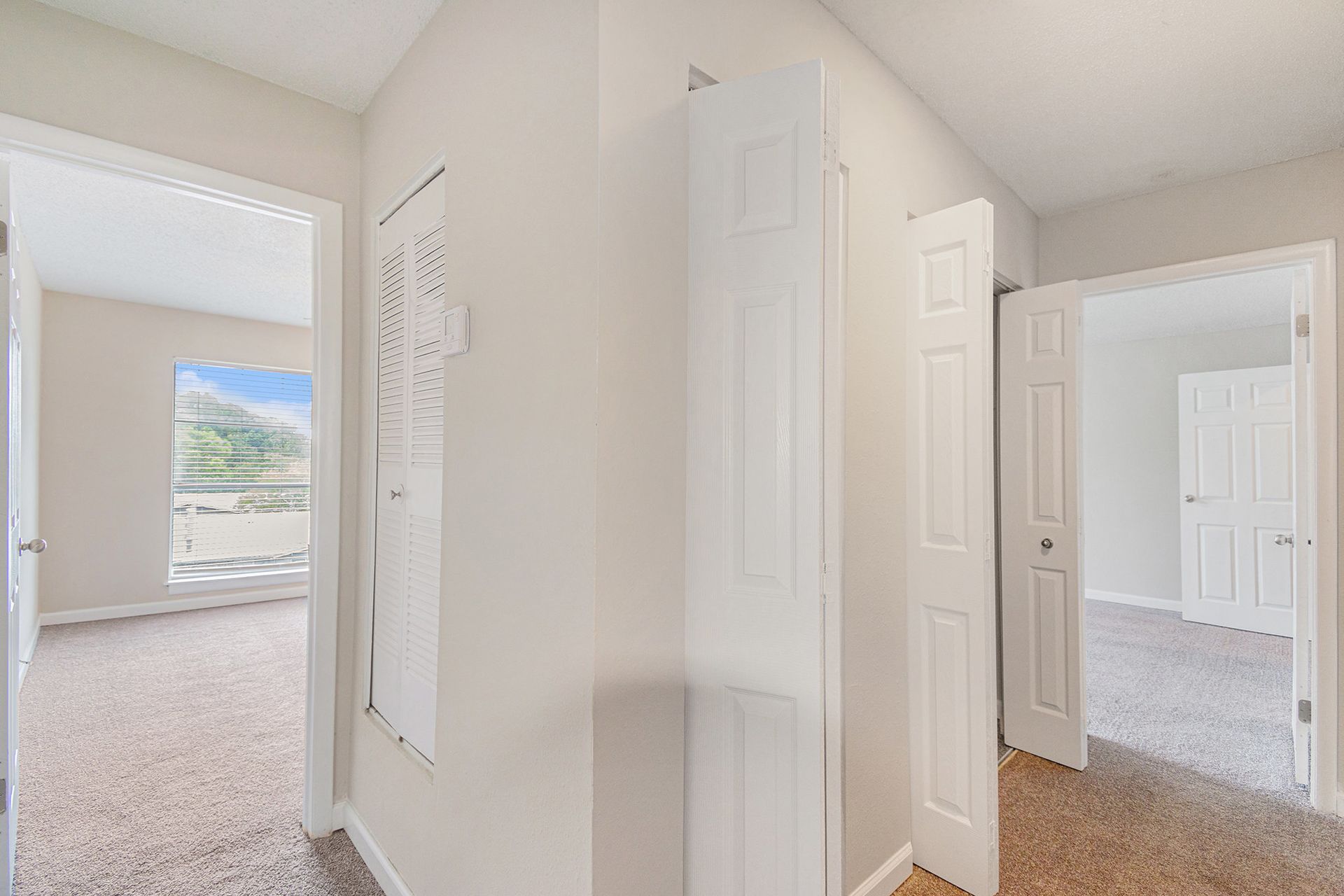 A hallway with light-colored walls and carpet, featuring white paneled doors and an open doorway to a bedroom with a window.