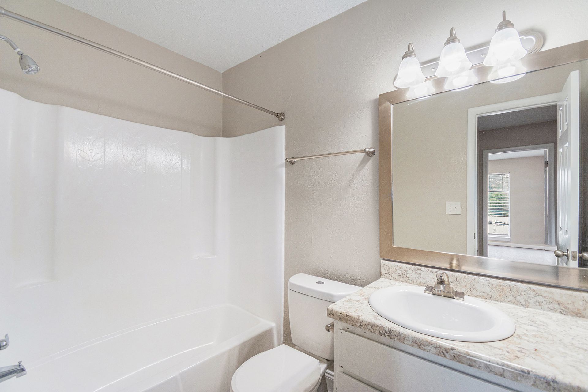 A bright, neutral-toned bathroom featuring a white bathtub, a toilet, and a vanity with a mirror and modern lighting.