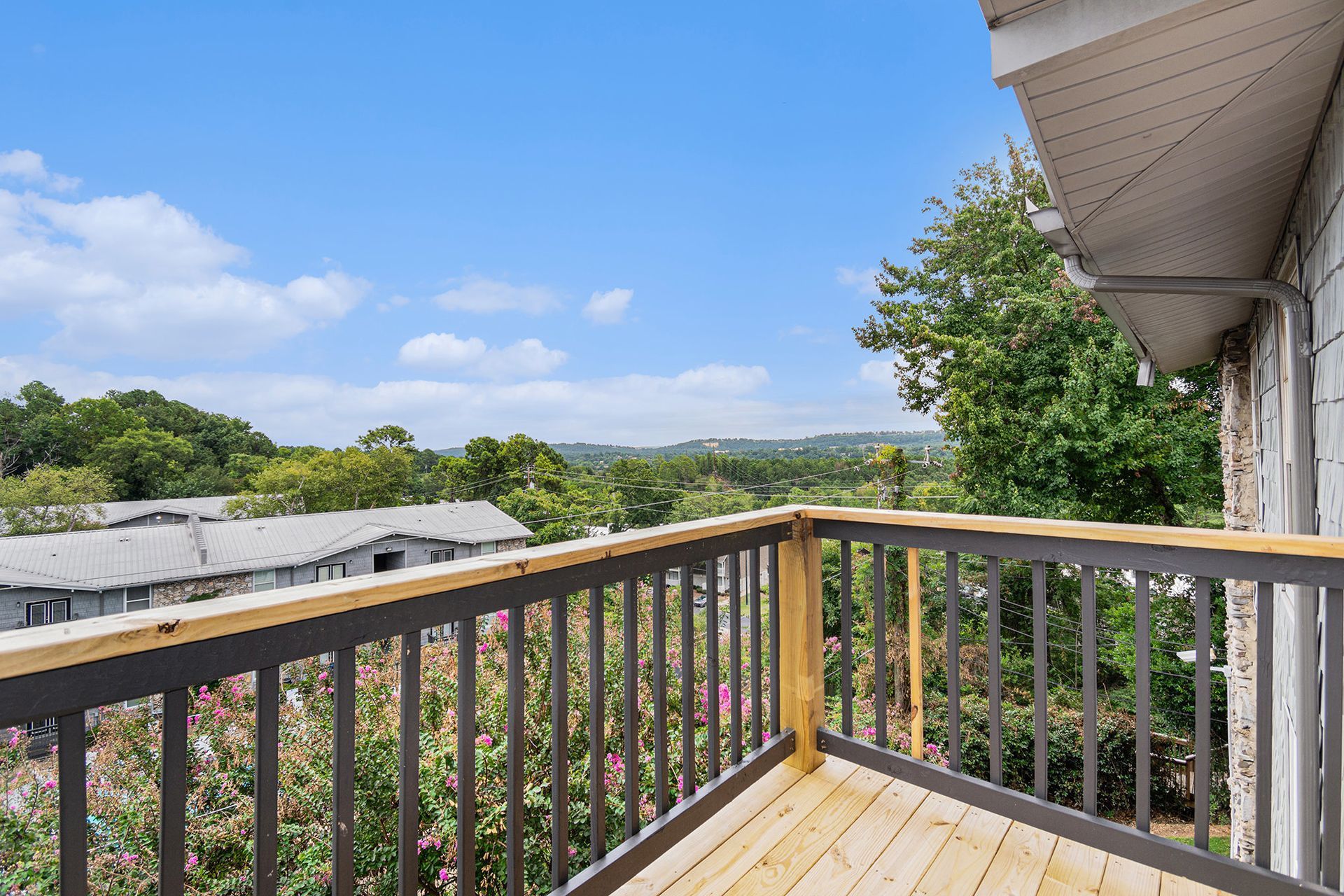 A wooden balcony with a grey railing overlooks a lush green landscape and a neighboring building under a blue sky.