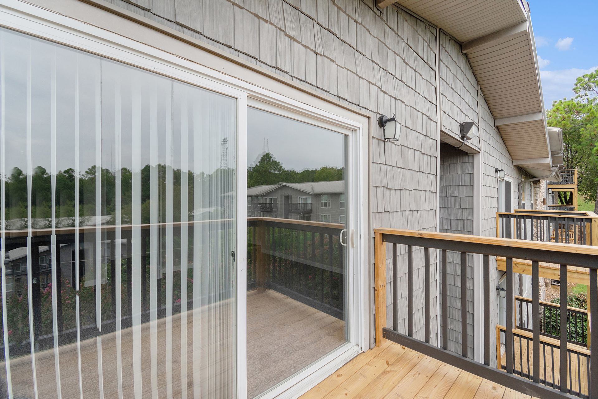 A wooden deck with a gray railing attached to a building featuring light gray wood siding and a glass sliding door.
