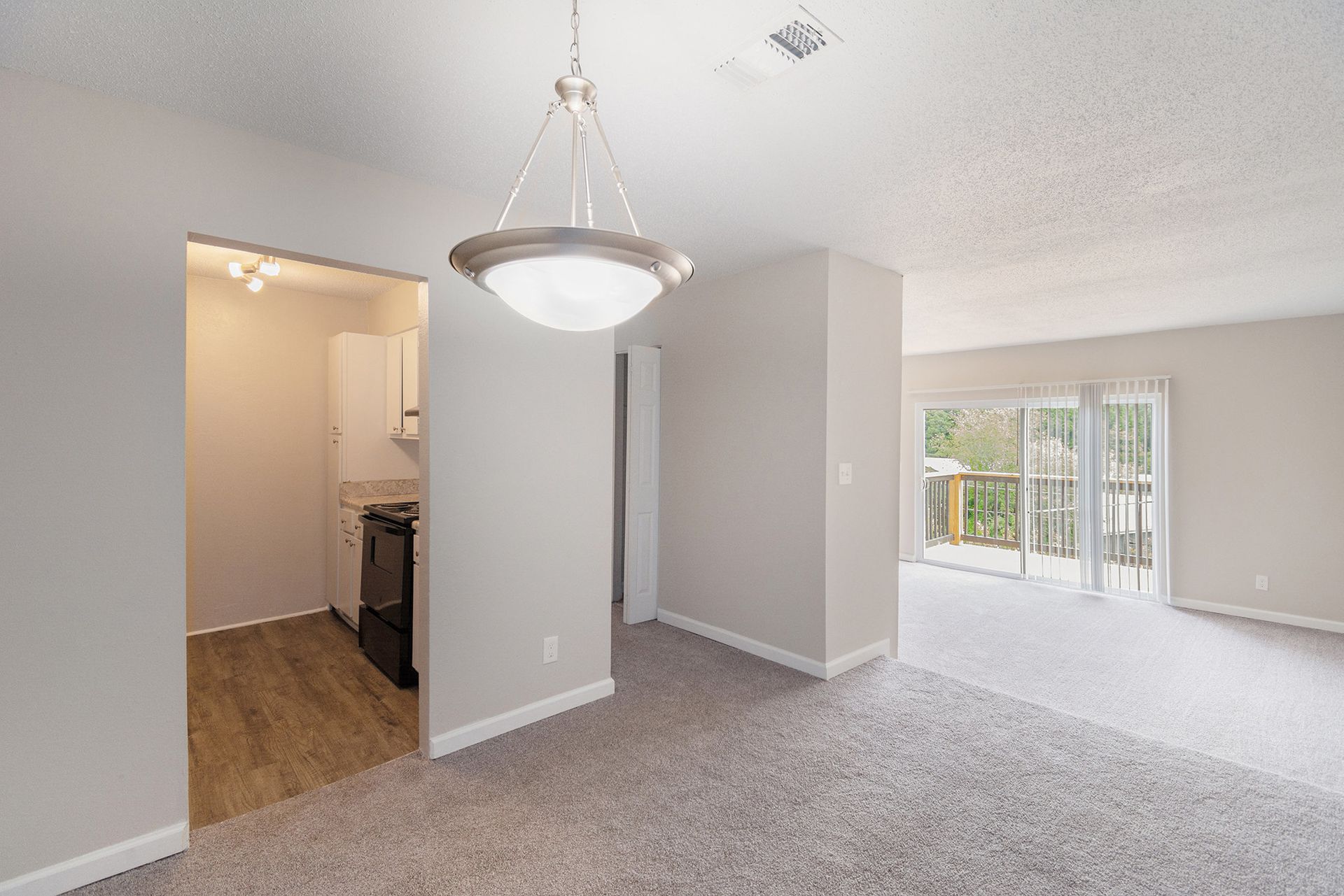An interior view of a dining area with a modern light fixture, connecting to a kitchen with wood floors and a living room.