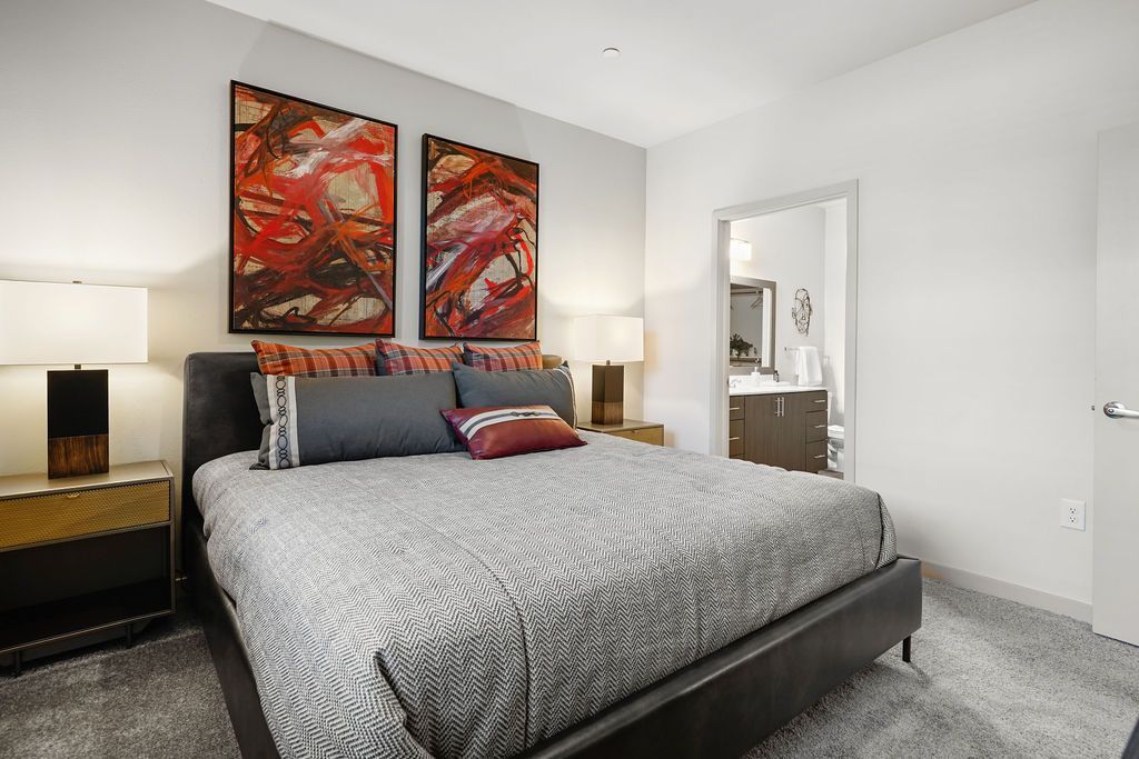 Bedroom in an apartment with a large bed, two nightstands, lamps, and an open bathroom doorway.