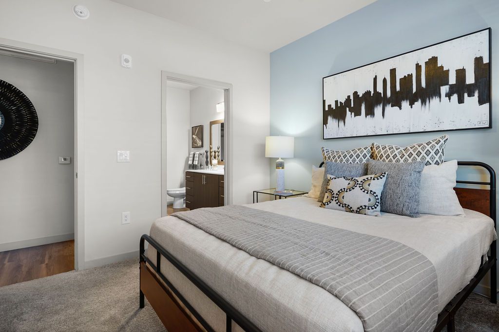 Bedroom in a modern apartment with a bed, throw pillows, nightstand lamp, and open bathroom door.