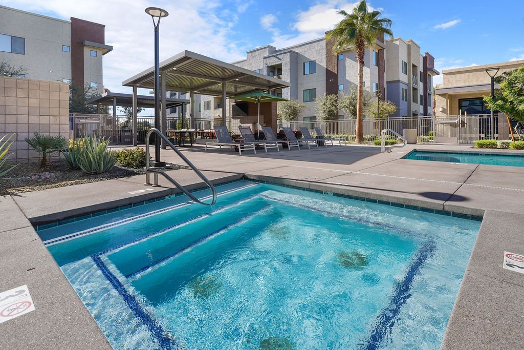 Outdoor pool area with a metal handrail, lounge chairs, shaded seating, and surrounding apartment buildings.