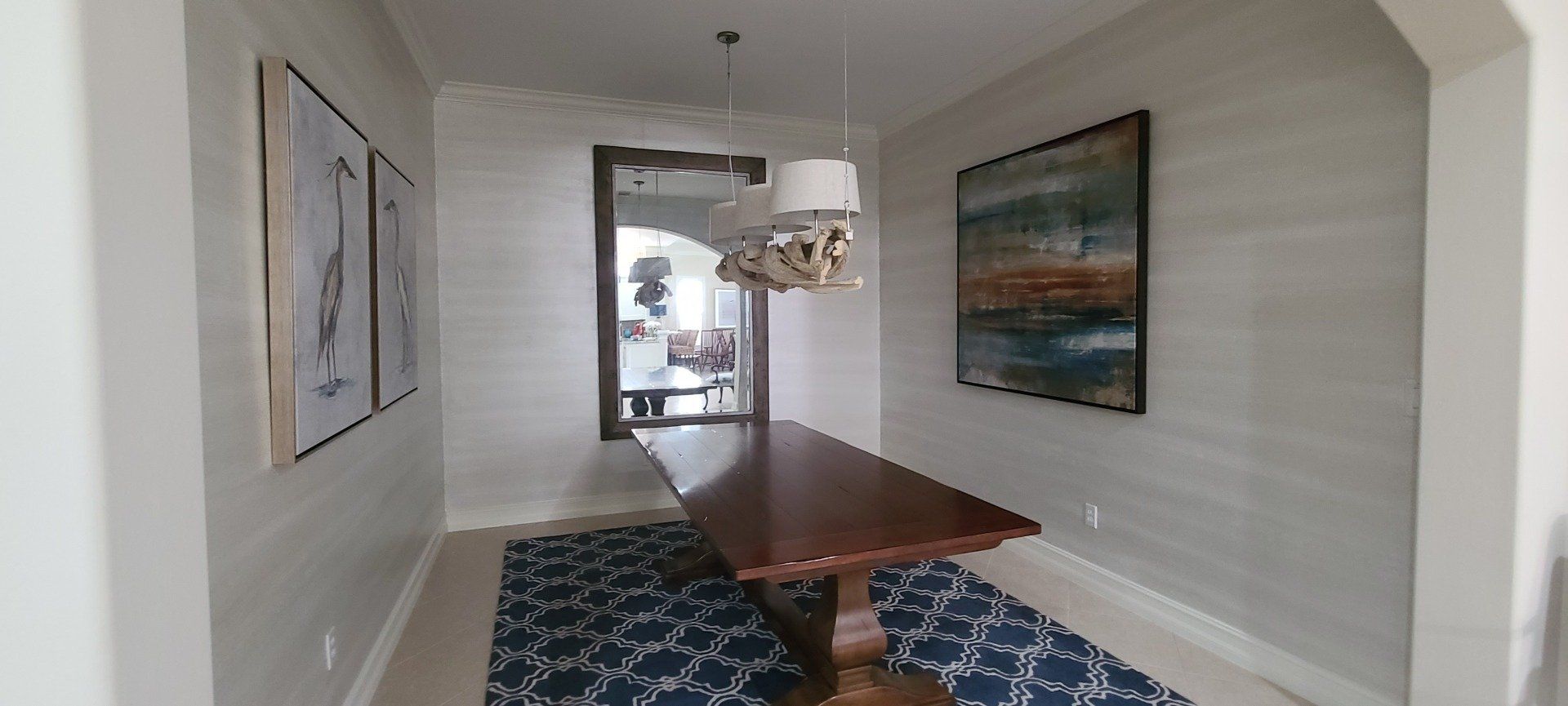 Dining room with long wooden table, blue patterned rug, framed art, large mirror.