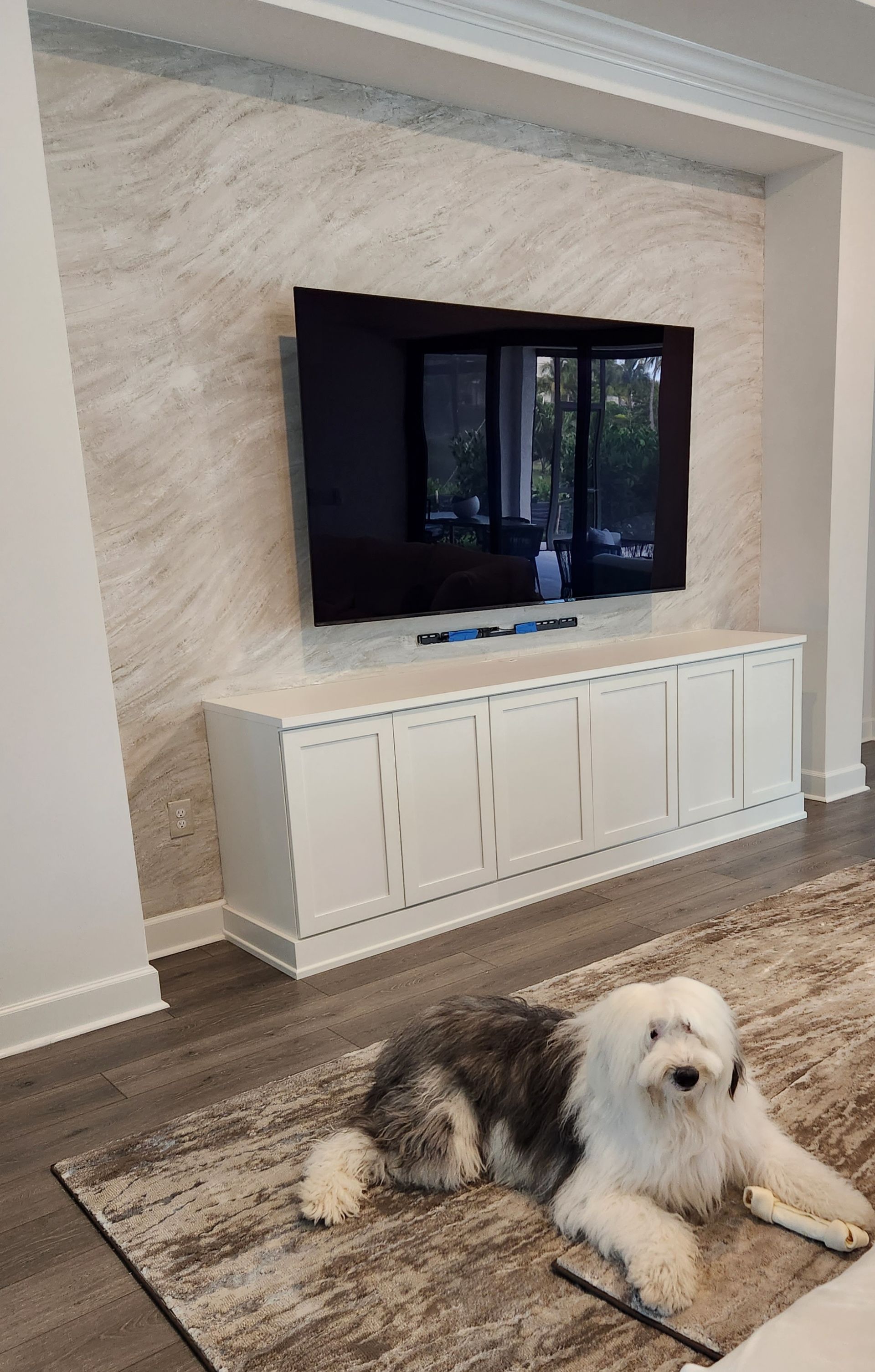 Dog lying on rug, looking at camera, below a TV mounted above a white cabinet against a stone wall.