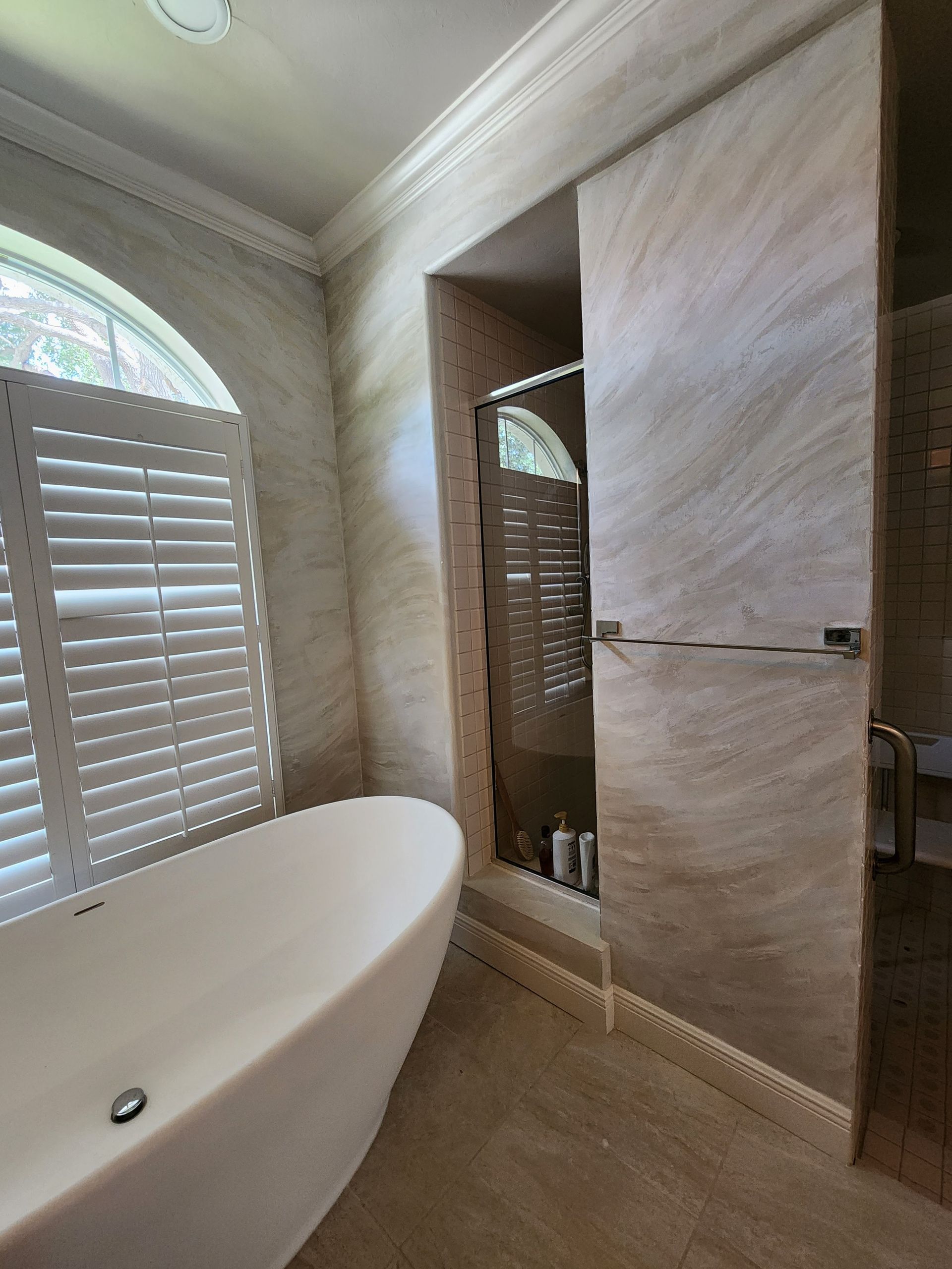 Elegant bathroom with a freestanding tub, shower, and shuttered window. Beige marble walls and flooring.