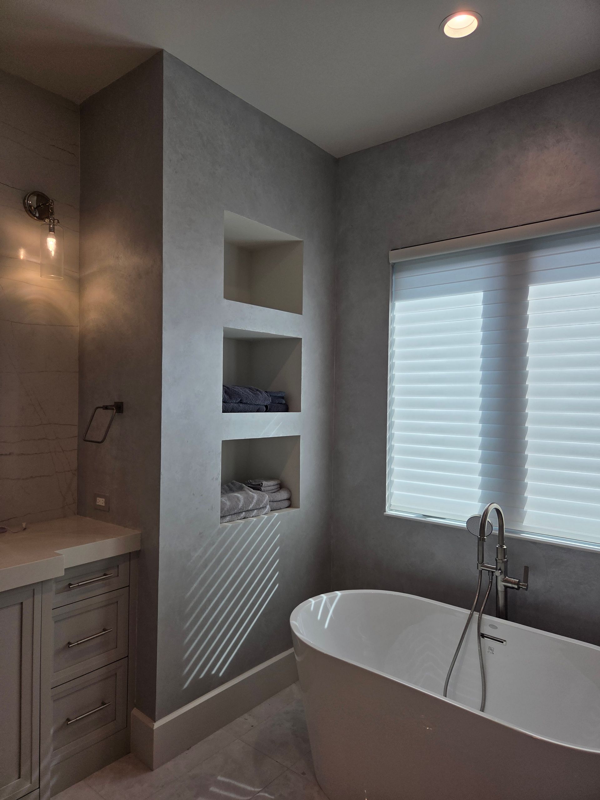 Modern bathroom with a bathtub, built-in shelves, and a window with blinds. Gray walls, white accents.