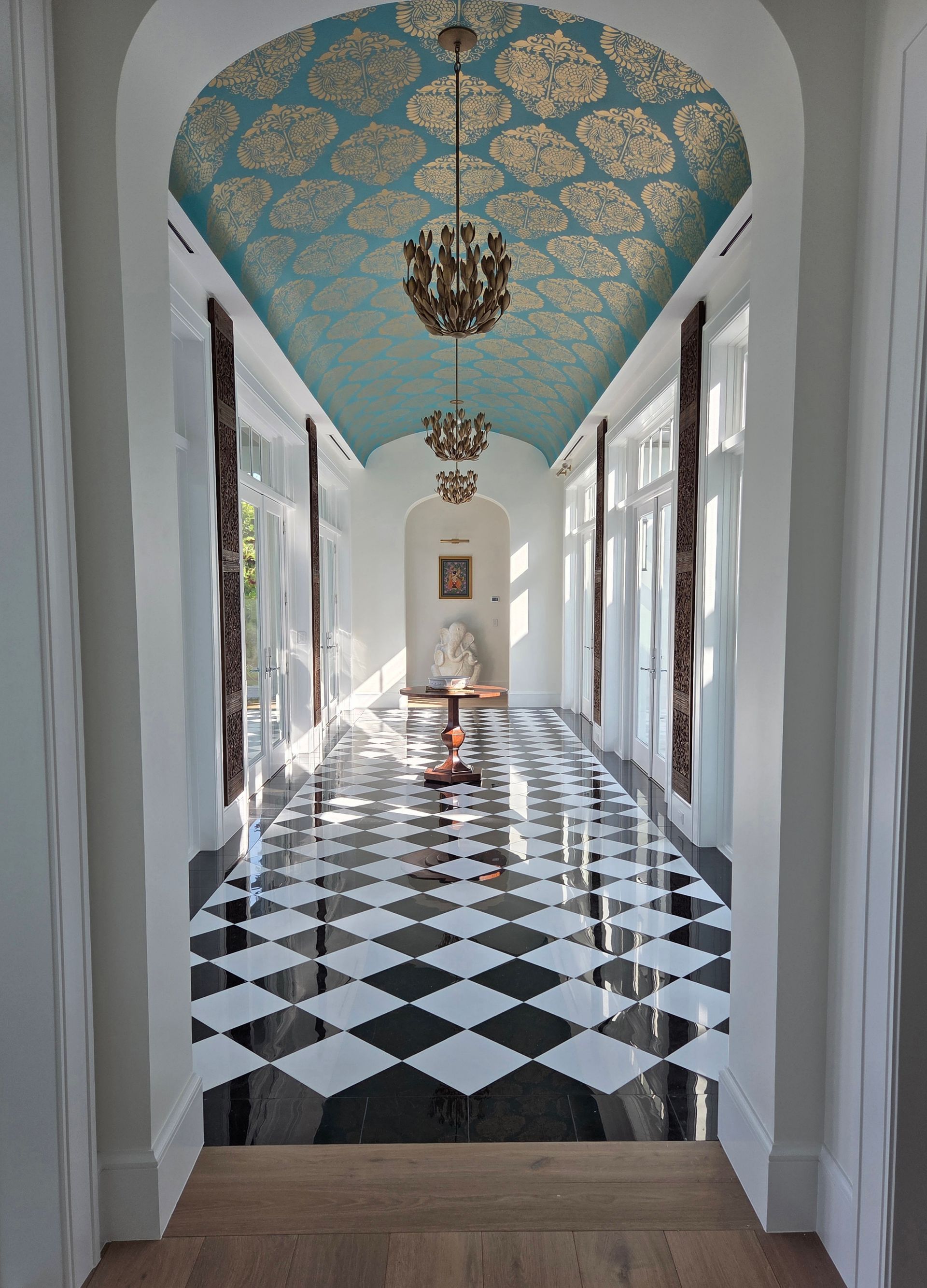 Long hallway with black and white diamond-patterned floor, ornate teal ceiling, and chandeliers.