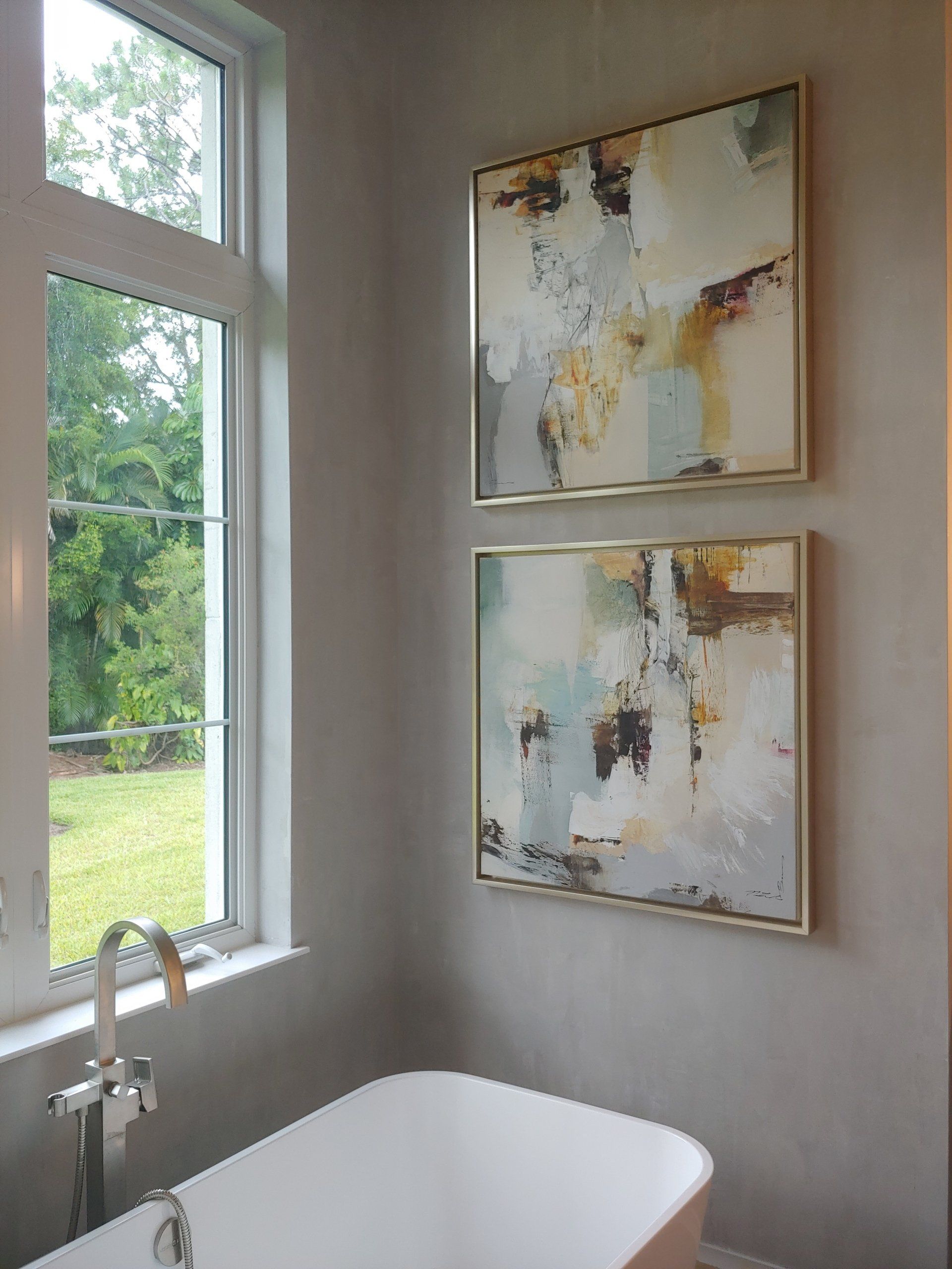 Two abstract paintings in gold frames above a bathtub near a window.