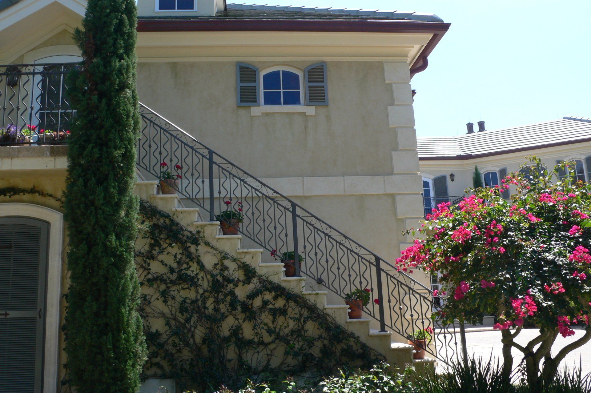 Exterior stairs with wrought iron railing, ascending to a cream-colored house with climbing vines and potted flowers.