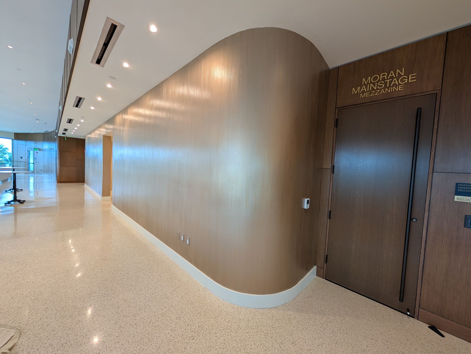 Hallway with curved, light brown wall, door to meeting room, speckled floor, recessed lighting.