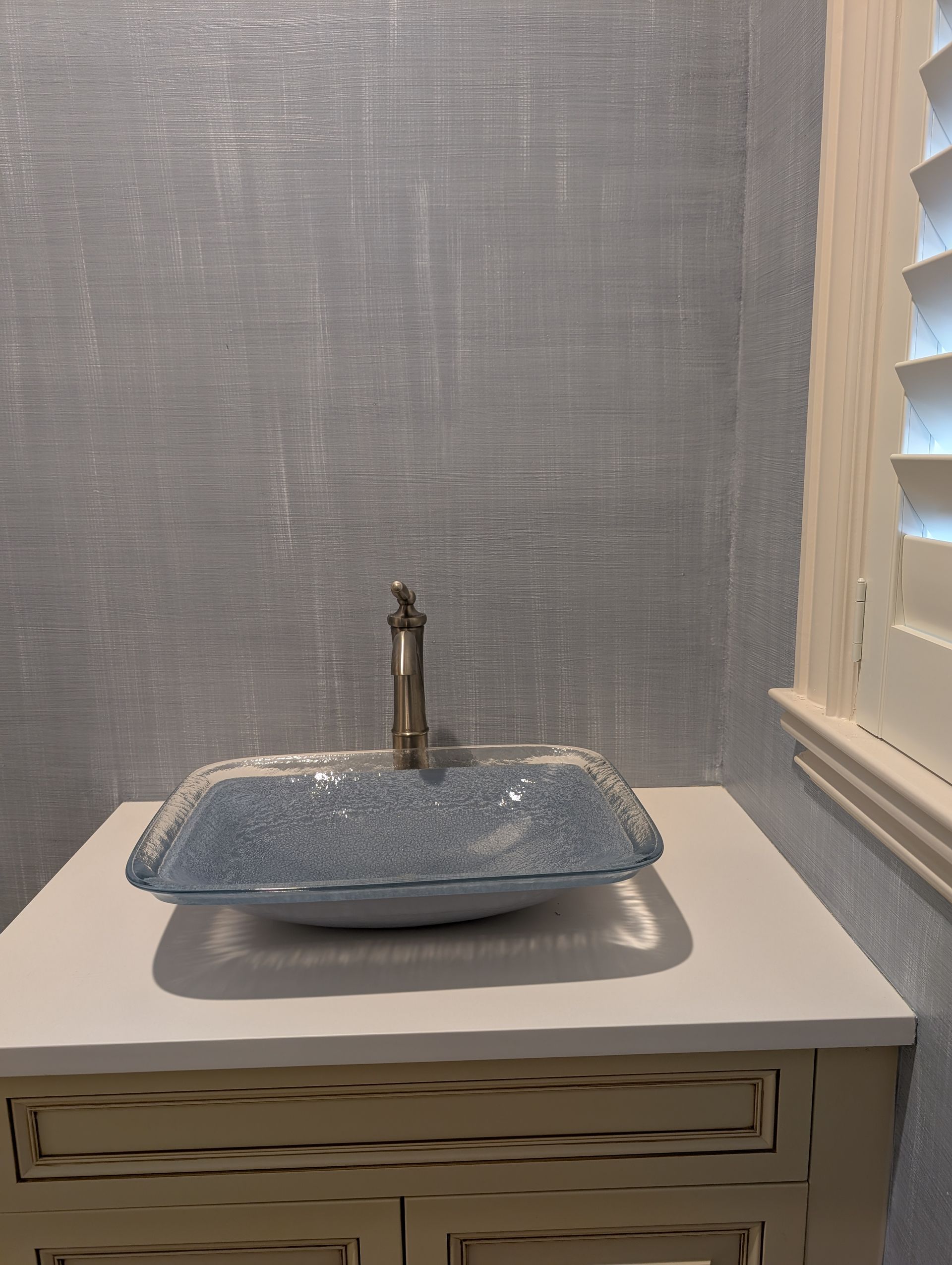 Blue vessel sink on a white cabinet, against a light blue textured wall. A window with white shutters is to the right.