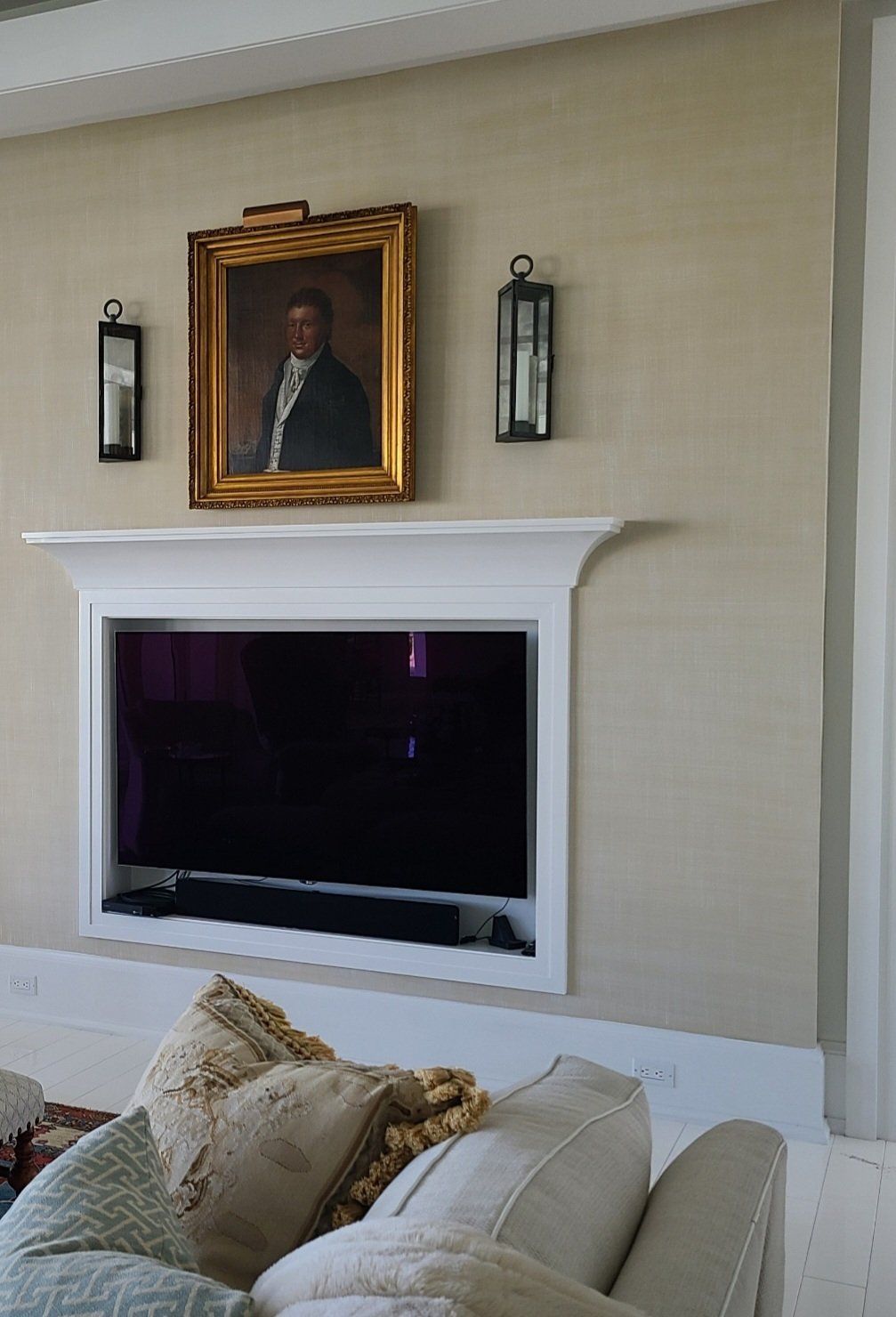 Wall-mounted TV above a couch, framed by a white mantel and a portrait, with sconces on a beige wall.