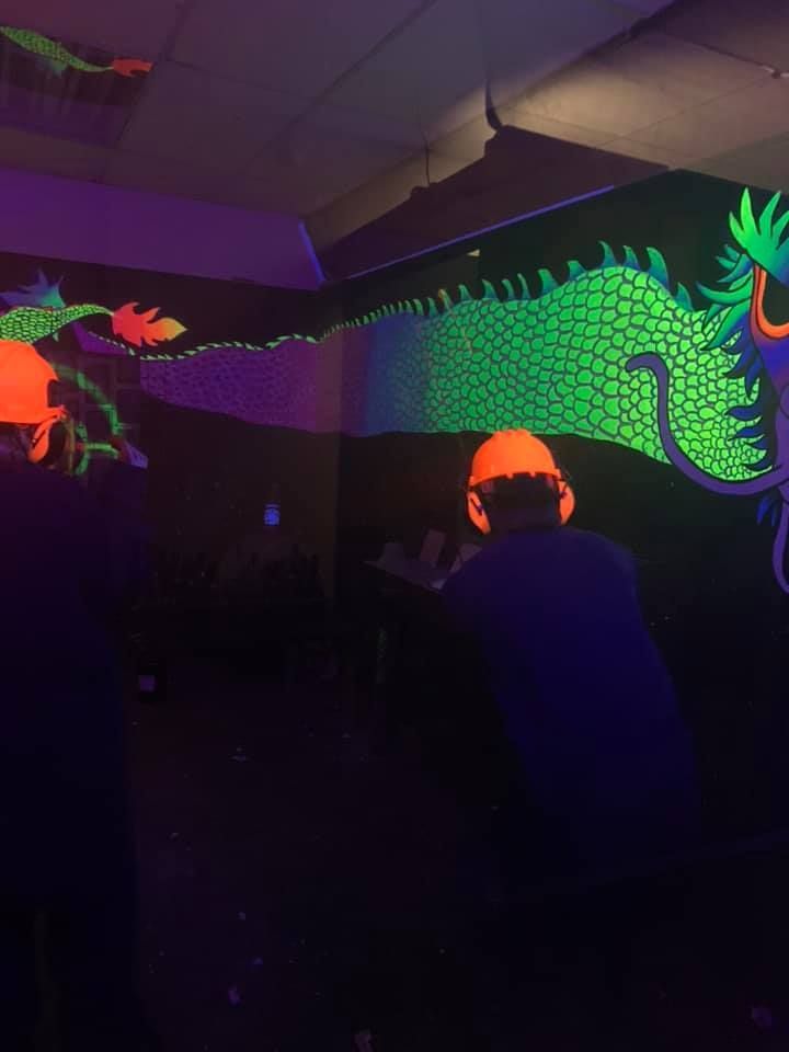 A man is standing in front of a glow in the dark dragon painting on a wall.