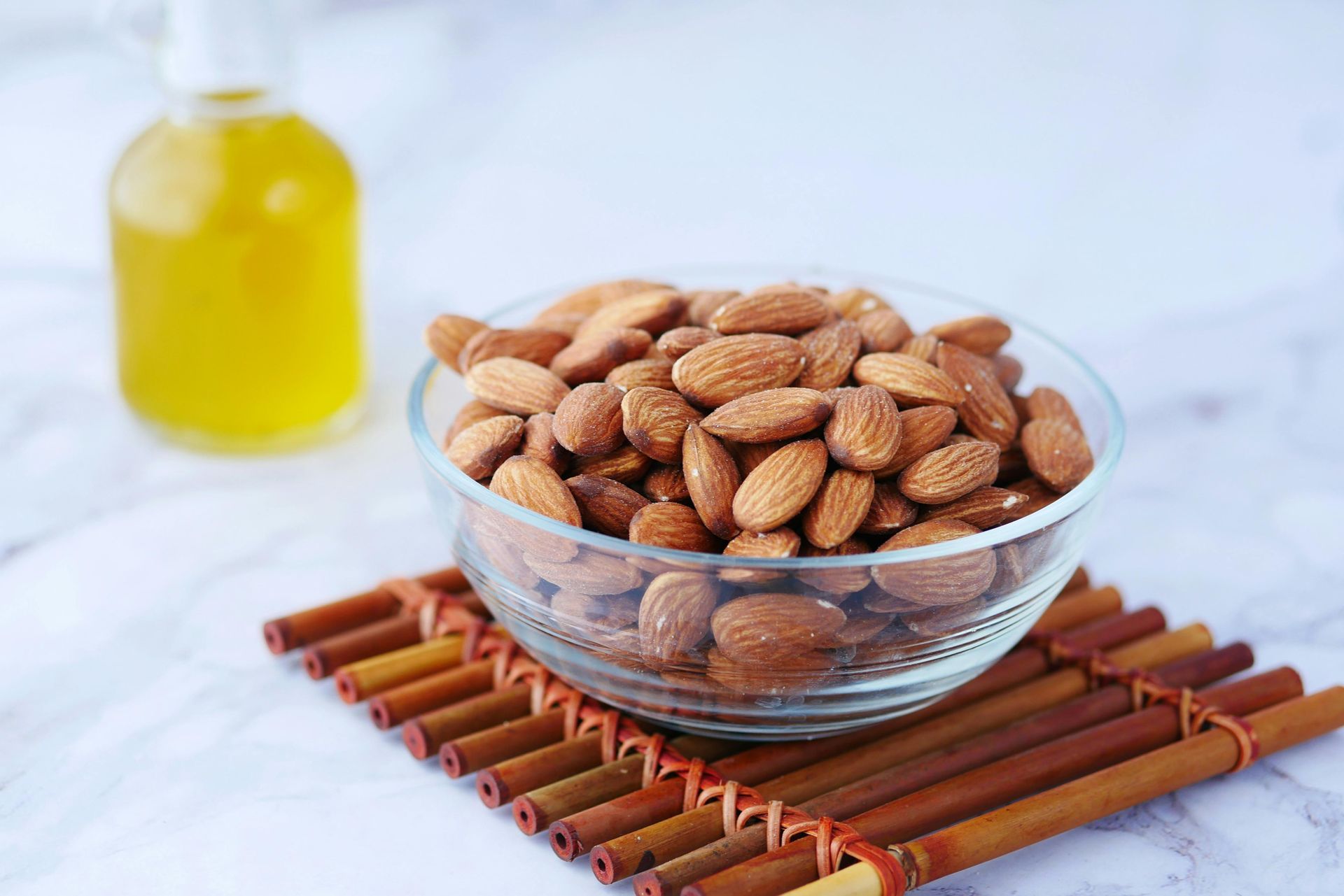 A glass bowl filled with whole almonds rests on a bamboo mat, next to a small bottle of yellow oil on a white surface.