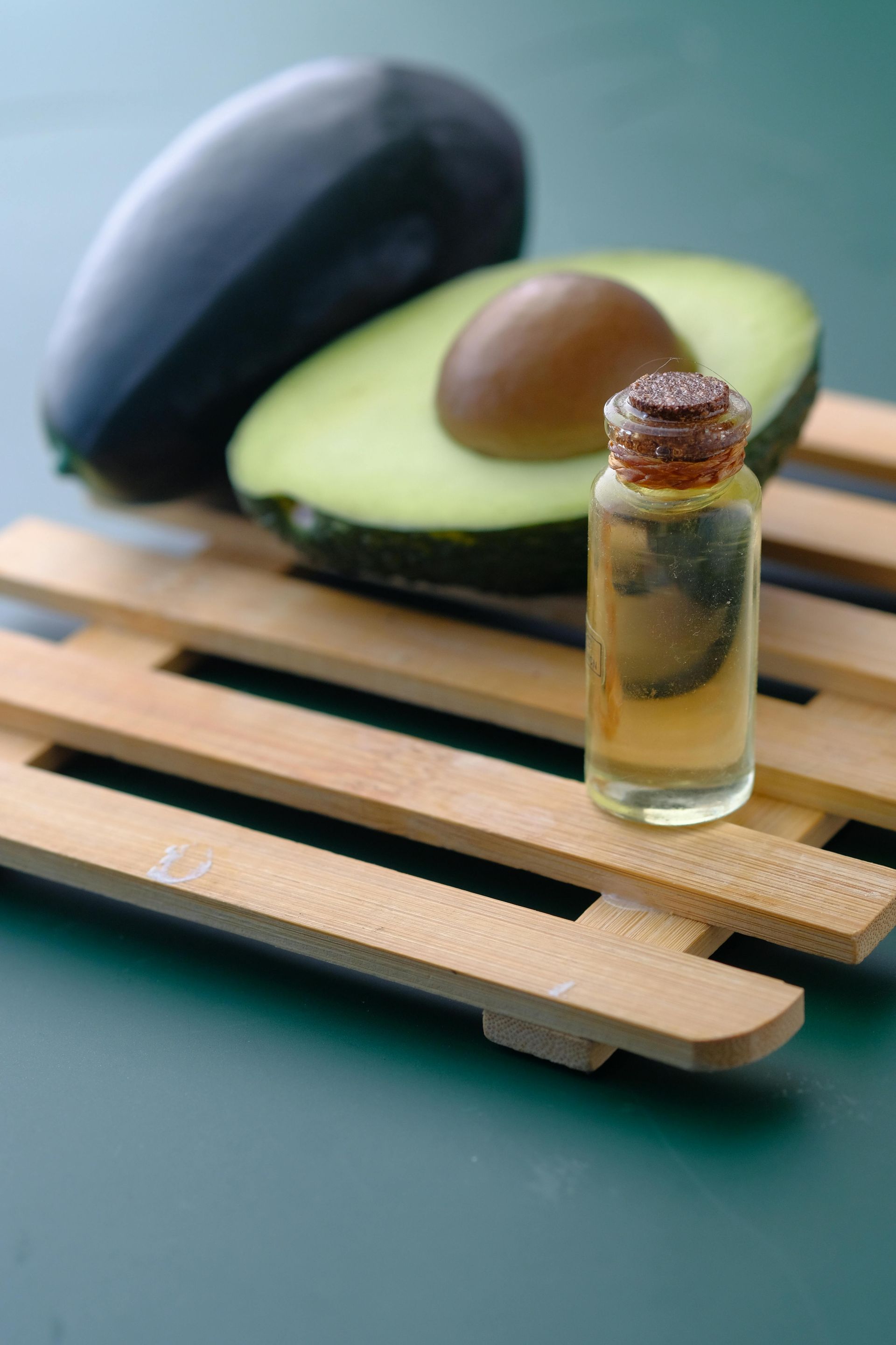 A whole avocado and a halved avocado with a pit sit next to a small bottle of golden oil on a wooden rack.