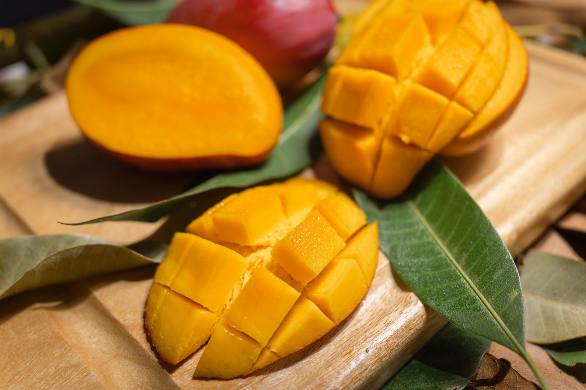 Sliced mangoes and a whole mango rest on a wooden cutting board, surrounded by green mango leaves.