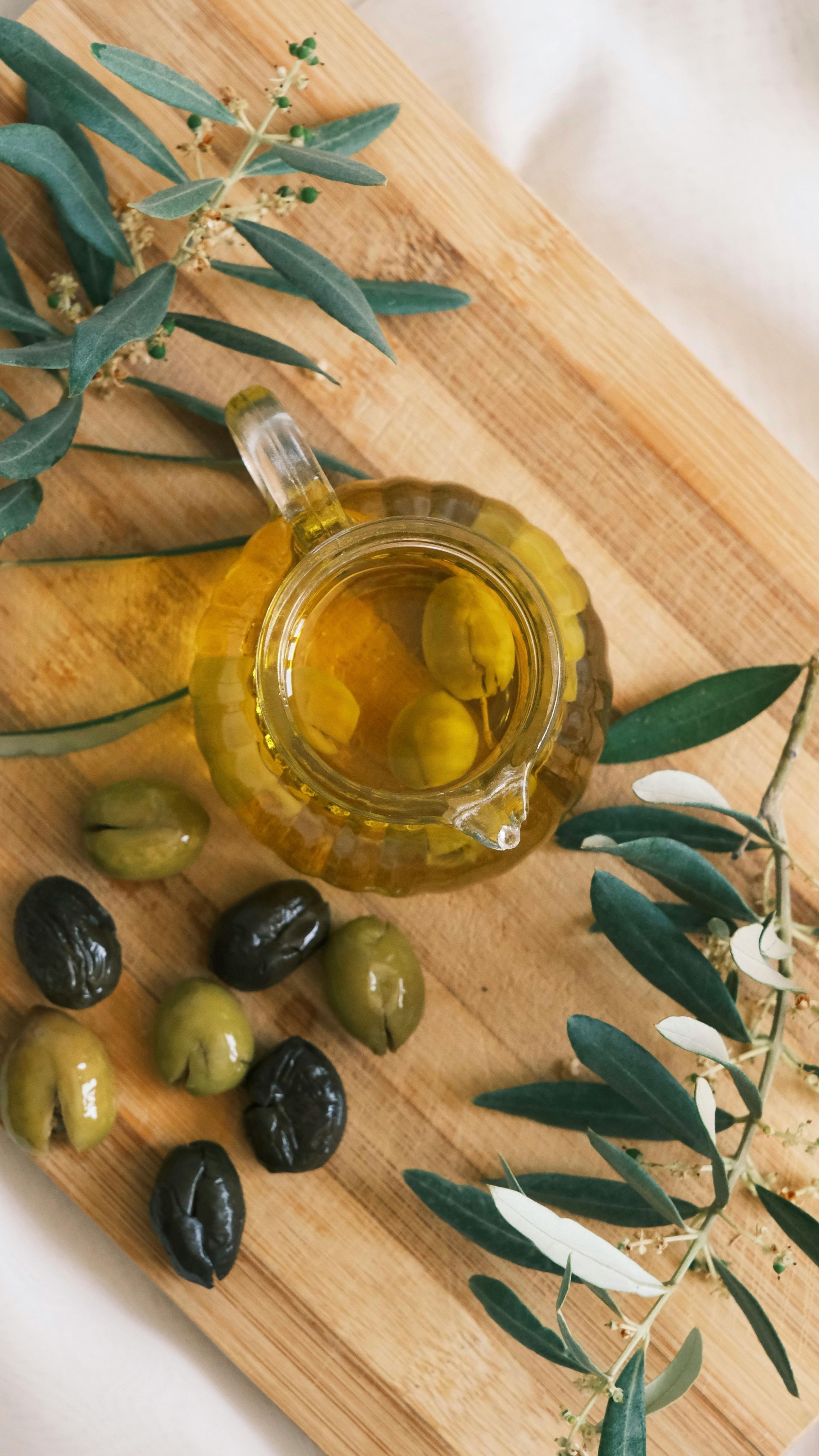 A glass carafe of golden olive oil sits on a wooden board surrounded by black and green olives and leafy olive branches.