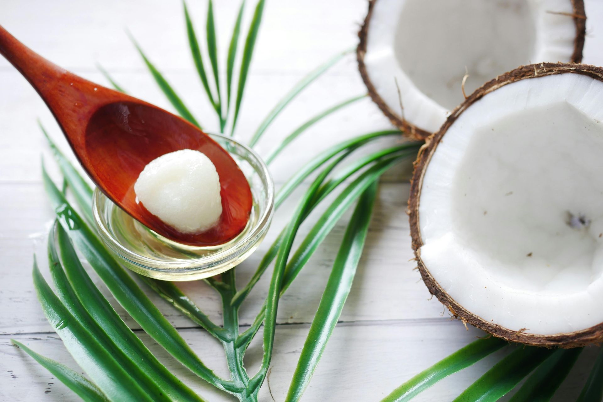 A wooden spoon holds a scoop of coconut oil over a glass bowl, with coconut halves and palm leaves on a white surface.