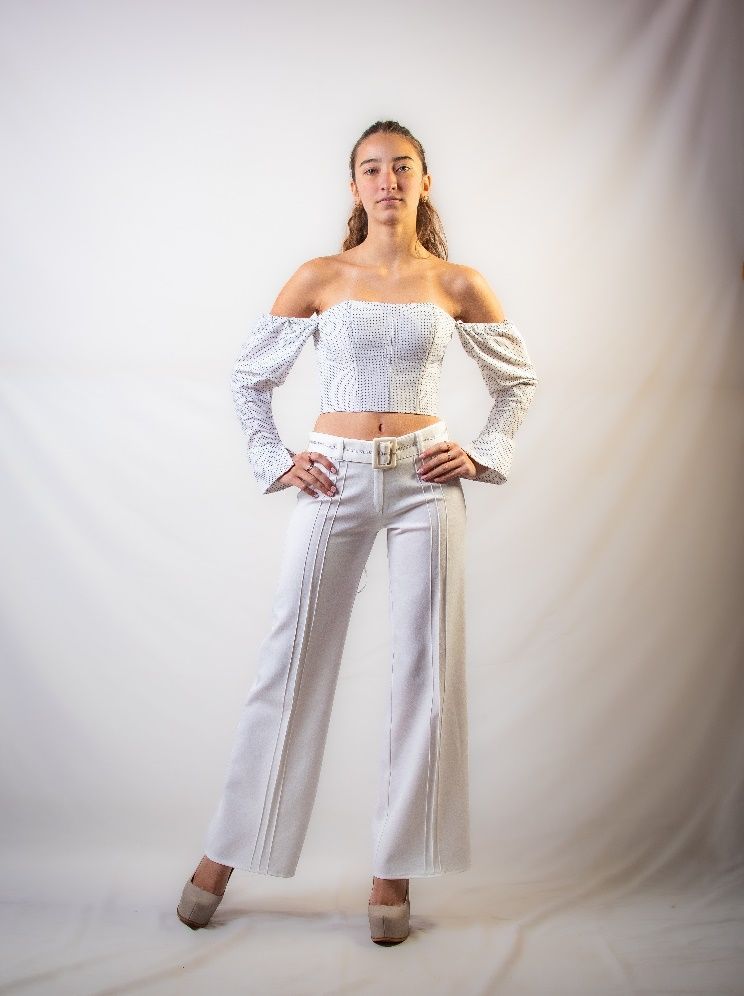 Una mujer con una blusa blanca con hombros descubiertos y pantalones blancos está de pie con las manos en las caderas.
