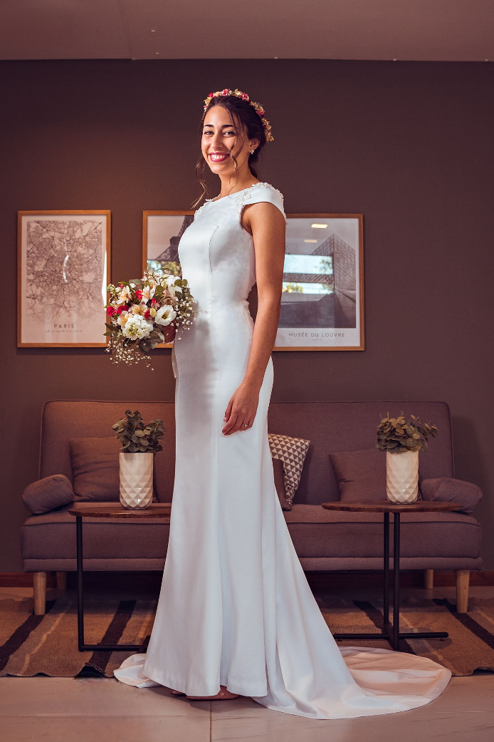 Una mujer con un vestido de novia está parada frente a un sofá sosteniendo un ramo de flores.