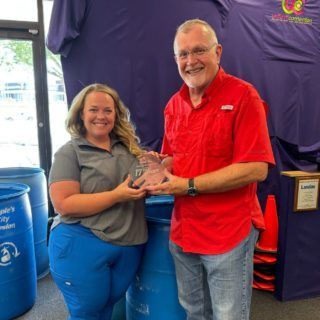 A woman and man hold an award. They stand by blue barrels and purple backdrop. The man wears red, smiling. The woman smiles too.