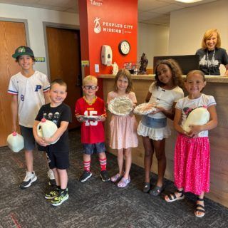 Children holding milk jugs and pizzas, smiling in a brightly lit office with a receptionist behind a counter.