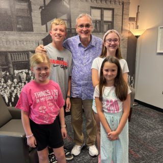A group of children and an older man pose indoors, smiling. They stand in front of a black and white mural.