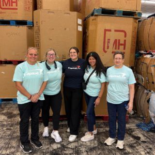 Five volunteers in teal shirts stand near boxes; warehouse setting.