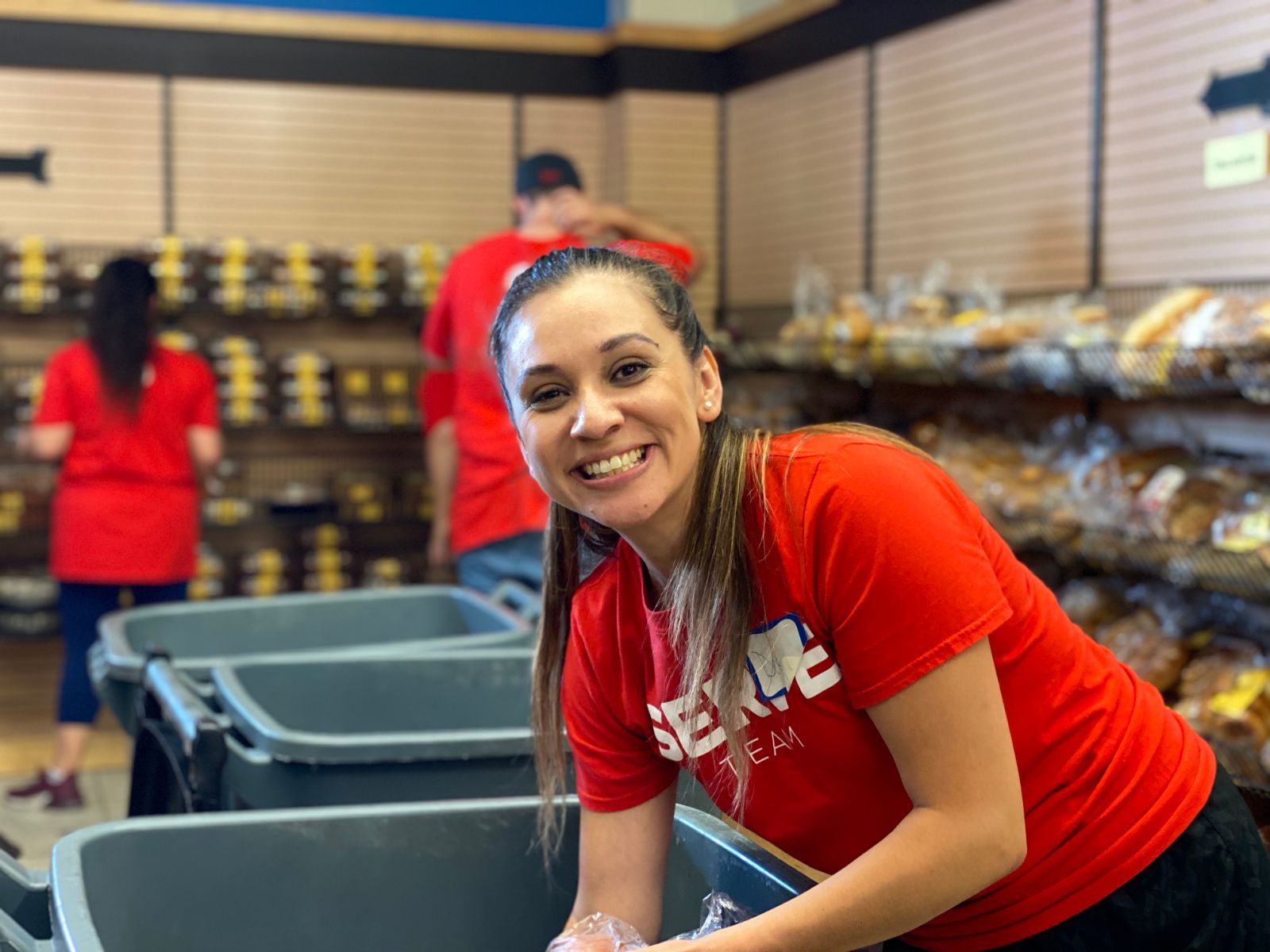 Woman in red shirt smiles at camera, working near bins in bakery with other volunteers.