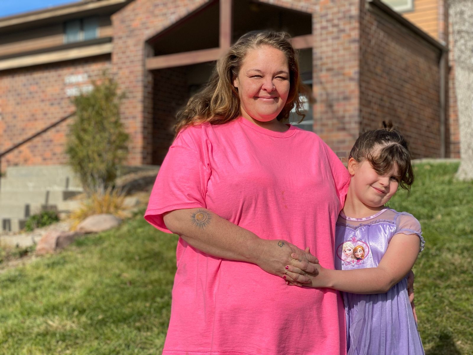 Woman in pink shirt embraces a young girl in a purple dress in front of a brick building.