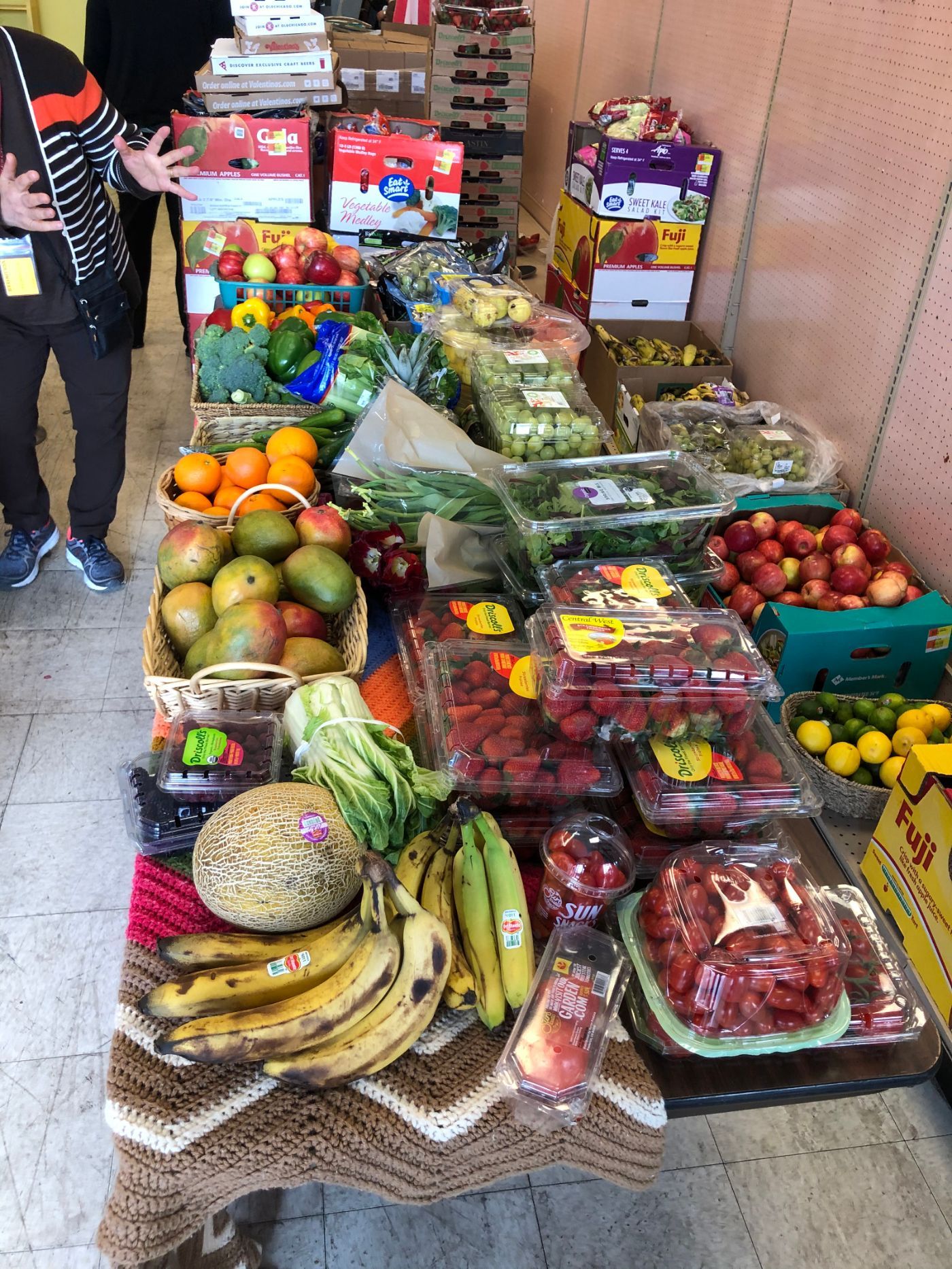 Produce table: mangoes, berries, lettuce, oranges, bananas, and more. People in background.