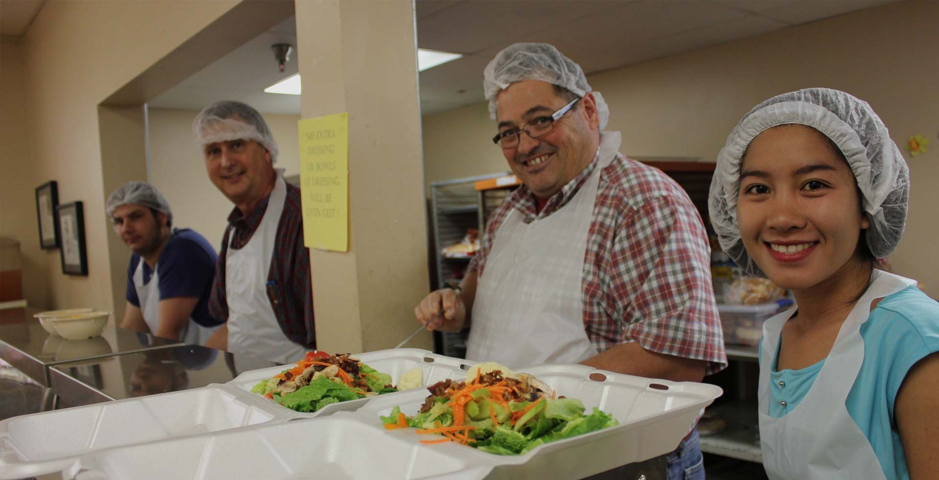 Volunteers preparing food in a kitchen, wearing hair nets and aprons. Smiling faces, two trays of food on a counter.