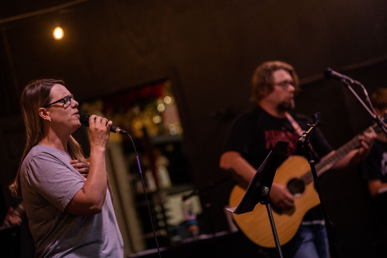 Woman singing into a microphone onstage, bandmate playing acoustic guitar in dimly lit venue.