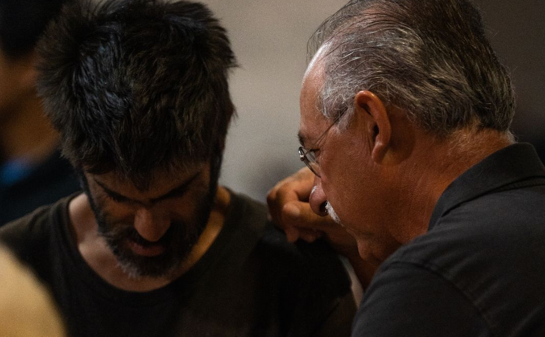 Two men in close conversation, one with dark hair looking down, the other older, wearing glasses, leaning in.