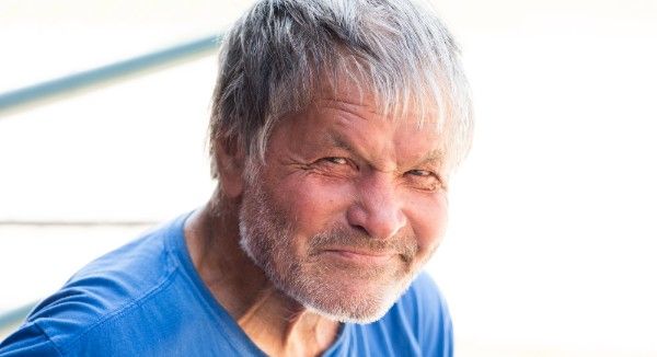 Older man with gray hair and beard in a blue shirt, squinting at the camera.