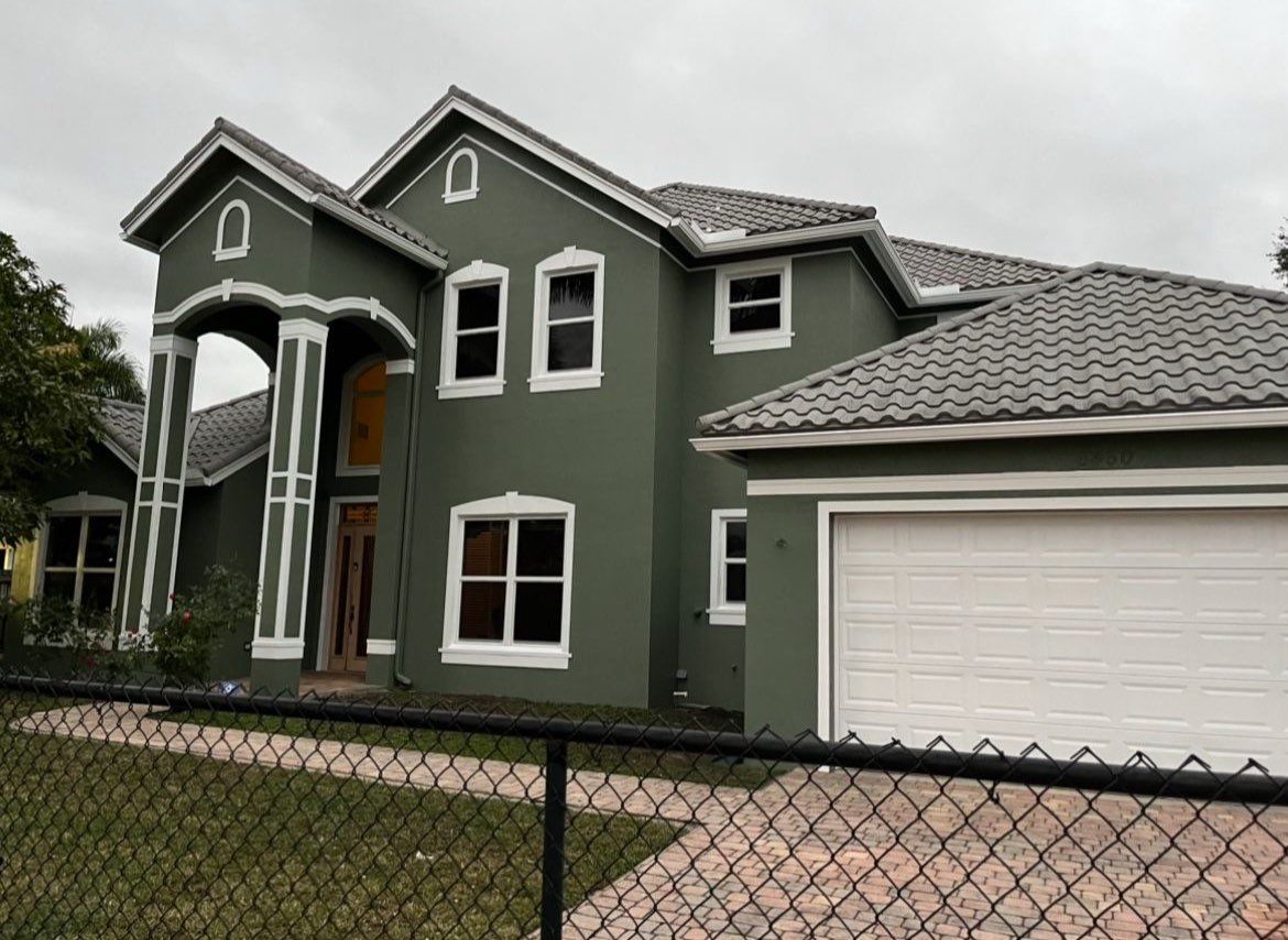 A large green house with a white garage door is behind a chain link fence.